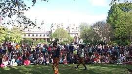 An outdoor crowd watches two performers. Behind them is an attractive white building with spires and a collonade.