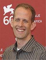 A blonde man is smiling. He wears a stripped shirt, and he is standing in front of a red wall.