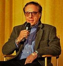 A white male in his 60s sitting in a chair while speaking into a microphone. He is wearing eyeglasses and a grey jacket over a blue buttoned down shirt. His left hand is rested on his lap.