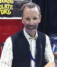 A middle-aged caucasoid man looking at the photographer; he is presented as a bust, wearing a yellow-plaid collared shirt under a cardigan sweater.