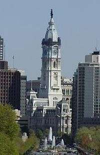 Distant ground-level view of a large building with a white exterior and a tall spire; the spire has a rounded roof and is topped with a black statue.