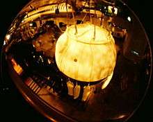 View from above of a large metal sphere suspended by ropes from a large hall's ceiling; 3 sand bags can be seen hanging on ropes under the sphere