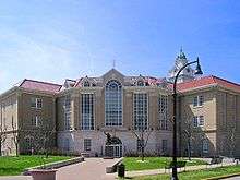 A large, brown building with towering windows, a clock tower, and a statue of a man in front