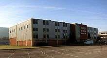 A white van is parked in front of a rectangular three-story building with many windows. A sign on the building says, "Piper Aviation Museum".