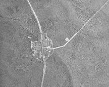 Black and white aerial view of a forested area with a vertical curving highway running through a group of large buildings near the center. A smaller road runs diagonally to the top right corner.