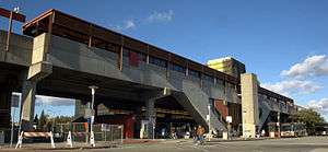 An elevated train station situates behind an empty road. Sections of the station is fenced and two buses wait on the side of the road.