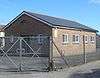 Three-quarter view of a pale brick hut with a shallow tiled roof, standing in asphalt-covered grounds behind a wire fence.  There is a recessed entrance door between two windows, and four windows in the longer side.