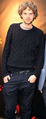 Three-quarter body shot of a 33-year-old man. He stands facing the camera with his right shoulder raised and hands placed into his jeans' pockets. He wears a dark jumper over a pale tee-shirt. His brown hair is slightly curly and about collar length. He has a slight beard.