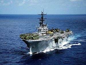 An amphibious assault ship sailing towards the camera. Multiple, large helicopters sit on the flight deck.