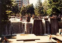 Columns with water flowing over the top, falling into a pool with square platforms. People are sitting and standing on the columns as well as the square platforms. In the background are trees and other buildings.