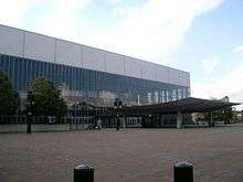 One side of a building mostly of glass windows, with a covered entrance extending to a brick plaza. Two trees grow alongside the building, and a partly cloudy sky appears in the background.