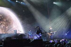 A stage shot of six men playing, at left is a man seated at keyboards with a microphone, next is the drummer behind his kit with a drumstick raised in his right hand, next is a guitarist singing into a microphone, then another singer-guitarist who is highlighted by overhead lighting, another man playing a bass guitar is mostly obscured behind the lead singer, the last man at right is also playing a guitar. In front of the stage are cameramen and audience members. Above is further stage lighting with, at left, a large projected image of the top part of the moon.