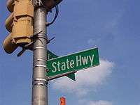 A green street sign reading State Highway attached to a traffic light pole.