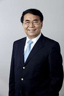 a smiling, aged man, wearing glasses, a suit and a blue tie while looking straight into the camera