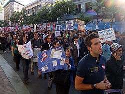 People peacefully marching up a street. Several are carrying signs displaying pro-same-sex marriage slogans, such as "We all deserve the freedom to marry." Most people have sad or serious facial expressions.