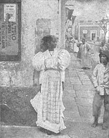 A dark-skinned woman in a light dress standing against a wall