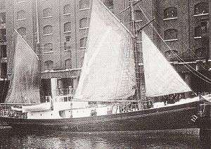  A ship with a black hull and white upper parts, three sails raised, in dock alongside a high multi-windowed warehouse building