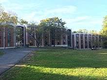 Pictured: Rothschild, Garrison, and Taylor Residence Halls (left to right) housed in one continuous, multi-level building. There are two visible entrances. The entrance connecting Rothschild and Garrison is one floor above the entrance connecting Garrison to Taylor, as the path along the building is at a slight incline. There are large rock formations visible in front of the building between the entrances, and medium-sized trees on the small patch of grass in front of the building.