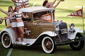 Six women, wearing a uniform of a white skirt and a white and gold cropped top with the word "Tech" on the front, ride onto the football field on the running boards and rear seat of a white-and-gold-painted antique car.