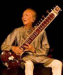 An old man sits on a platform and holds a long-necked lute while looking to the side