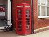 A red, cast iron telephone kiosk with a domed roof. it stands against a red brick wall at the corner of a building.