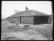 Man standing on roof of dilapidated sod house with axe or similar implement