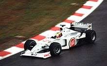 A figure, wearing a helmet of a dark blue design with red and yellow stripes, is driving a Formula One with an white colour scheme adorned with sponsorship. He is holding both hands on a steering wheel, turning right.