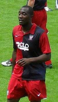 A man with bald hair who is wearing a red and blue striped top and red shorts. He is standing on a grass field.