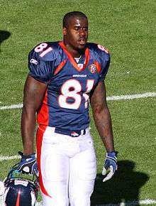 In the foreground, an American football player holding a blue helmet, blue jersey (number 81), and white pants walks along the sideline.