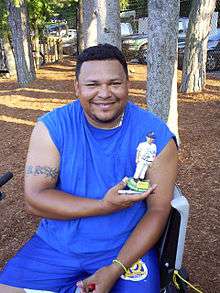 A man wearing a blue sleeveless T-shirt and blue shorts smiles while holding a bobblehead depicting himself in a white baseball uniform.