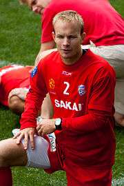 A blond Caucasian man wearing red football kit with his right knee raised.