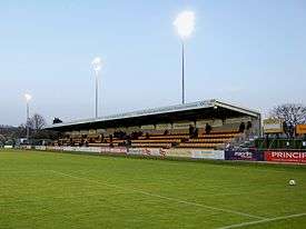 A single-tiered stand which has nine rows of alternating gold and black seats. In front of the stand is a field of grass upon which a solitary football sits.