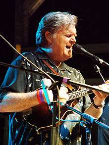 A man with white hair playing a guitar in front of two microphones