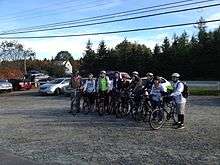 A photograph of twelve people standing in a row wearing bicycles and straddling or standing next to bicycles, all looking left under a blue sky