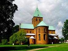 A cruciform, copper-roofed, brick church in Romanesque style with rounded window arches and domed apse. The large tower has pointed Gothic-styled windows.