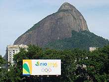 An advertising sign appears in front of some trees. Buildings are visible behind the trees as well as a large rocky hill in the background.