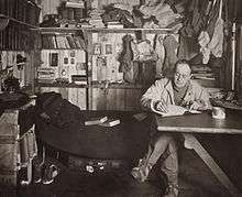 Man sitting cross-legged at table, pipe in hand, apparently writing. Much clutter of clothing, books and equipment is in the background.