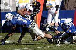 An American football player in a blue jersey tackles a player in a white jersey on a football field.