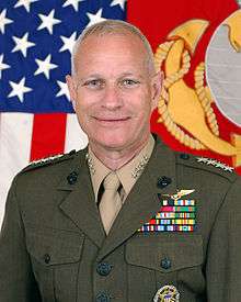 A color image of Robert Magnus, a white male in his Marine Corps Service A uniform. He is not wearing a hat, several ribbons are visible as well as a round badge and naval aviator insignia. The Marine Corps flag and United States flag are visible in the background.