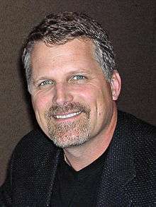 A man with grey hair, wearing a black a suit.