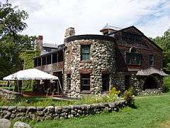 A mansion made of stone, brownstone, and wood framing sheathed in cedar shakes. A porch and patio are to the left of a two story turret that centers the image, and the patio is protected by a white pavilion tent.