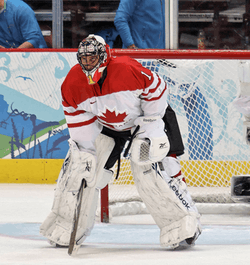 An ice hockey goaltender wearing a red mask, white pads and a white and red jersey with a maple leaf logo.  He is bent over with his hands at his knees and his head looking forward.