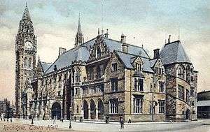 A large, ornate and angular taupe-coloured building on a sunny day. The building fills the image with the exception of flat, grey pavement at the building's frontage, which is dotted with people. The multi-storey building has many small slitted triangular and rectangular bays and windows across its façade, except for a dark arched entrance at ground-level. At the far end of the building is a huge clock tower topped with a trigangular spire standing tall at around fifteen to twenty times the height of an average adult human.