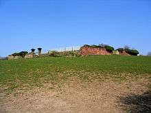 A ruined sandstone wall seen in the distance across a field, including a pair of gateposts on the left.