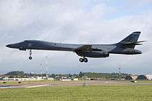 Gray aircraft before landing, flying left, with gears extended. Green grass make up the foreground. Buildings and communication towers are in the background