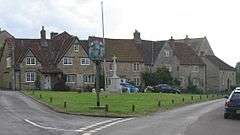 Street scene. Triangular area of grass with village sign on wooden post and stone cross behind. Stone houses with tiled roofs in the background.