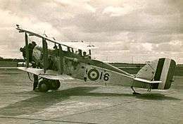 Manned single-engined military biplane parked on airfield, with propeller spinning