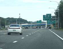 Ground-level view of four lanes of a busy freeway; several green exit signs and two overpass bridges are visible in the distance.