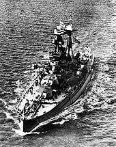 Aerial photograph of Royal Oak under way at sea, flying both the Norwegian flag and the White Ensign at half-mast.