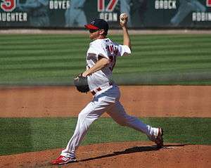 A man in a white baseball uniform and navy-blue baseball cap with a red brim throws a baseball with his right hand from atop a dirt mound on a grass field